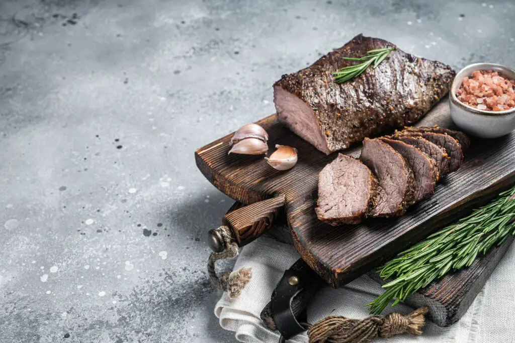 Roast and sliced tri tip beef steak on a wooden board with herbs. Gray background